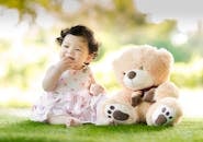 pexels-photo-1166473-1166473 Smiling baby girl sits on grass with a plush teddy bear, enjoying a sunny day.